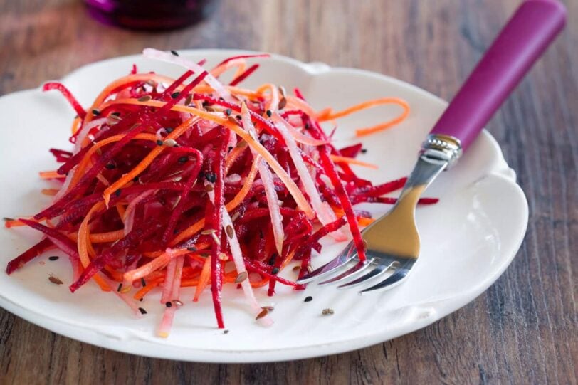 white plate on a wood table with beet, carrot and kohlrabi slaw on it next to a purple handled fork