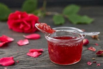 rose petal jelly in a jar with a spoon on a wooden table surrounded by rose petals