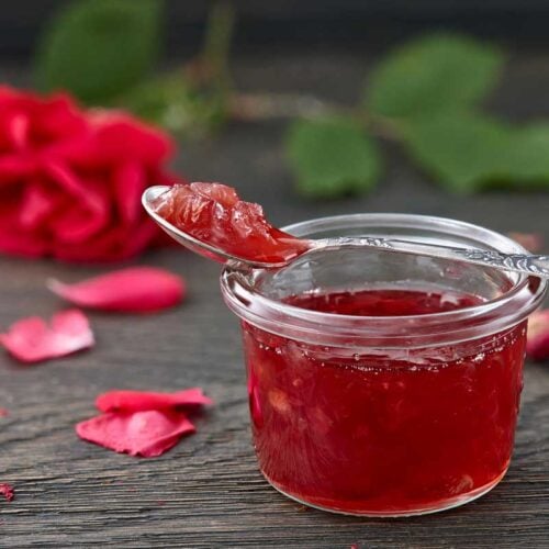 rose petal jelly in a jar with a spoon on a wooden table surrounded by rose petals