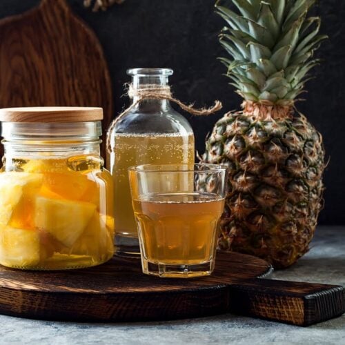 glass of tepache on a cutting board next to a bottles of fermenting pineapple