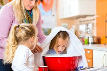 mother and daughter helping other daughter hold a towel over her head to inhale steam from a red bowl
