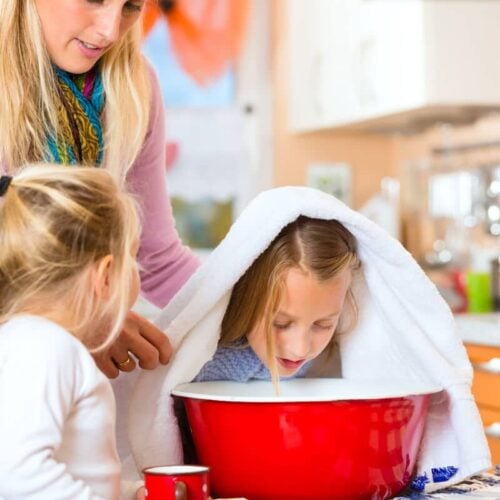 mother and daughter helping other daughter hold a towel over her head to inhale steam from a red bowl