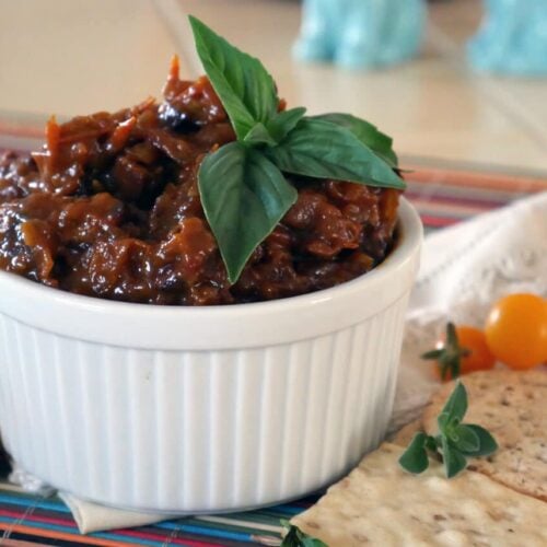tomato bacon jam in a white dish next to crackers and cherry tomatoes on a countertop.